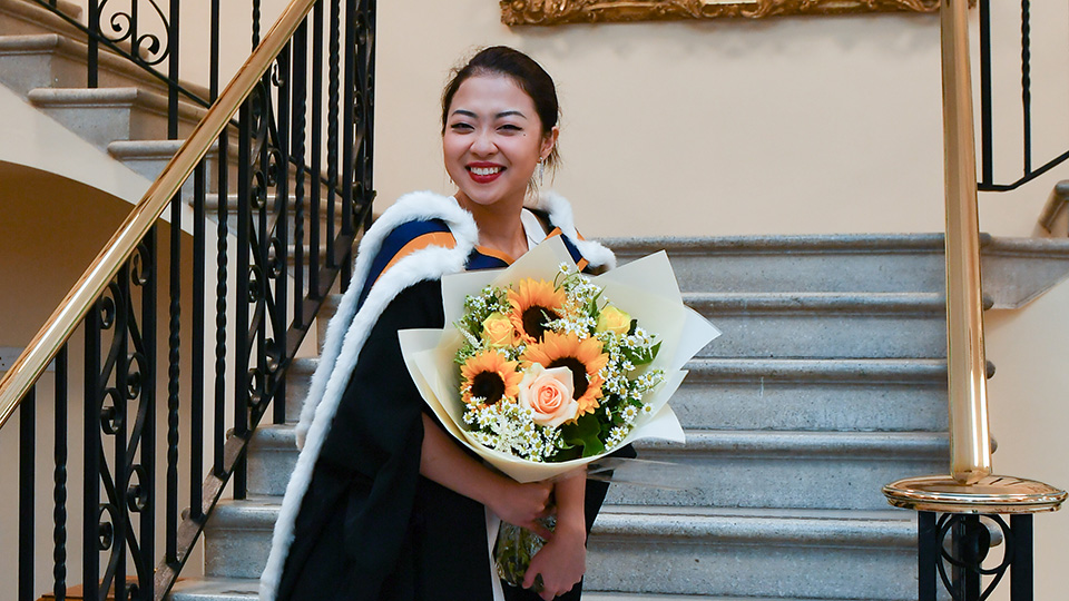 A graduate holding flowers and smiling, stood on steps at the RCM